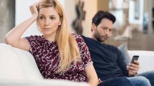 Woman sitting on couch looking distressed while her partner sits behind her on his phone, illustrating emotional distance in marriage.