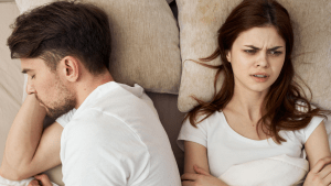 Wife lying awake with folded arms and an upset expression while her husband rests beside her, representing emotional distance in marriage