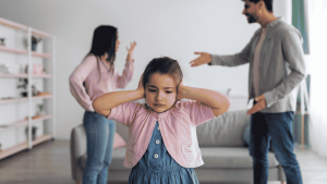 Little girl covering her ears while her parents argue in the background, representing how childhood experiences shape emotional safety in marriage
